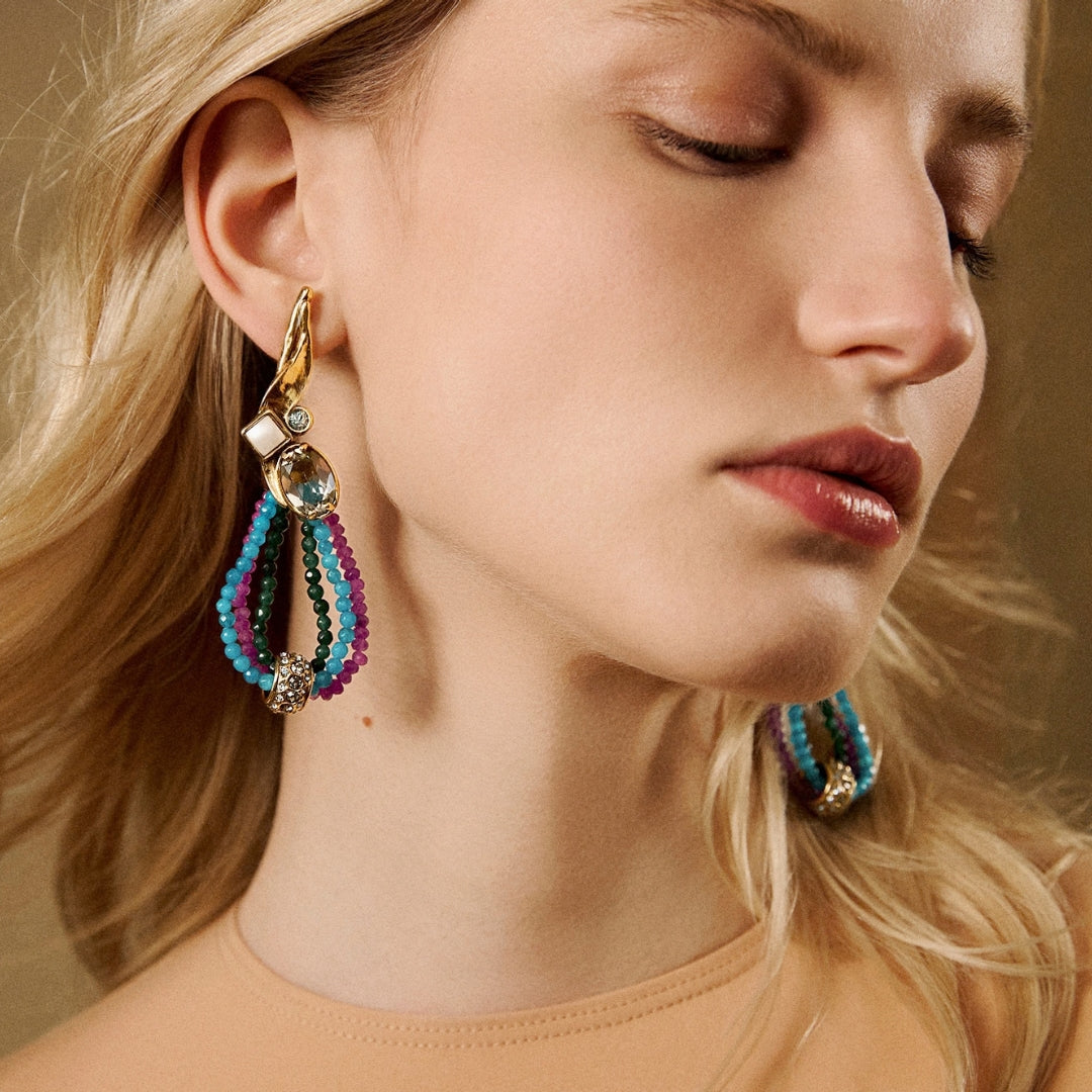 Brinco Camila Klein com design sofisticado e vibrante, inspirado nas cores do verão. A peça combina pedras naturais em tons de roxo, azul e verde, dispostas em delicadas formas esféricas e fios coloridos. O detalhe metálico dourado valoriza a composição, criando um acessório autoral, leve e cheio de personalidade para produções elegantes e contemporâneas.