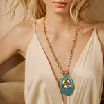 Colar Camila Klein com corrente dourada e pingente oval em pedra natural azul, com detalhe central em metal dourado. A peça combina cores e texturas orgânicas em um design autoral, sofisticado e vibrante, inspirado na leveza do verão. Ideal para compor produções elegantes e cheias de personalidade, valorizando a beleza singular das pedras naturais.