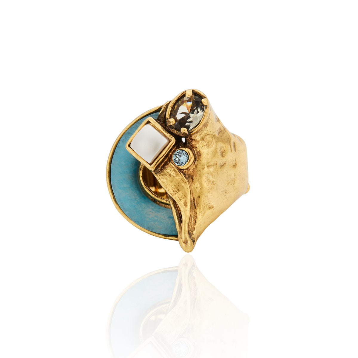 Broche da Coleção Ofertar, com design orgânico e sofisticado, acabamento dourado e composição marcada pelo contraste de cores e texturas. A peça combina elemento oval em tom verde profundo, cristais e pedras em diferentes formatos, incluindo detalhe quadrado branco e pedra azulada. Uma joia singular, inspirada no gesto de oferecer, com brilho, transparências e formas que celebram afeto e presença.